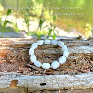 May include: A white gemstone bracelet with silver beads. The bracelet is made of smooth, rectangular stones and is lying on a piece of weathered wood.