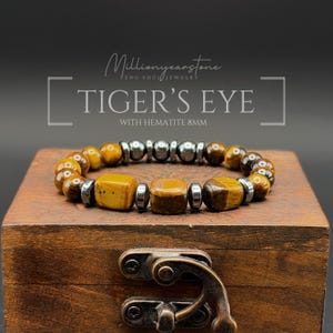 May include: A beaded bracelet featuring 8mm tiger's eye and hematite stones. The design includes round and square-shaped tiger's eye beads interspersed with metallic grey hematite spacers. Text on the image reads Millionyearstone TIGER'S EYE WITH HEMATITE 8MM.