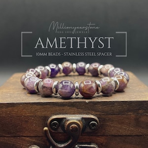 May include: Amethyst bead bracelet with 10mm beads and stainless steel spacers. The bracelet is displayed on a wooden box. The text on the image reads "Millionyearstone Feng Shui Jewelry" and "Amethyst 10mm Beads - Stainless Steel Spacer."