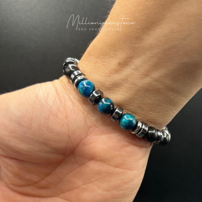 May include: A beaded bracelet featuring blue and black beads. The bracelet has a mix of round and cylindrical beads, with a metallic finish. The bracelet is worn on a wrist. The text "Millionyearstone FENG SHUI JEWELRY" is in the upper left corner.