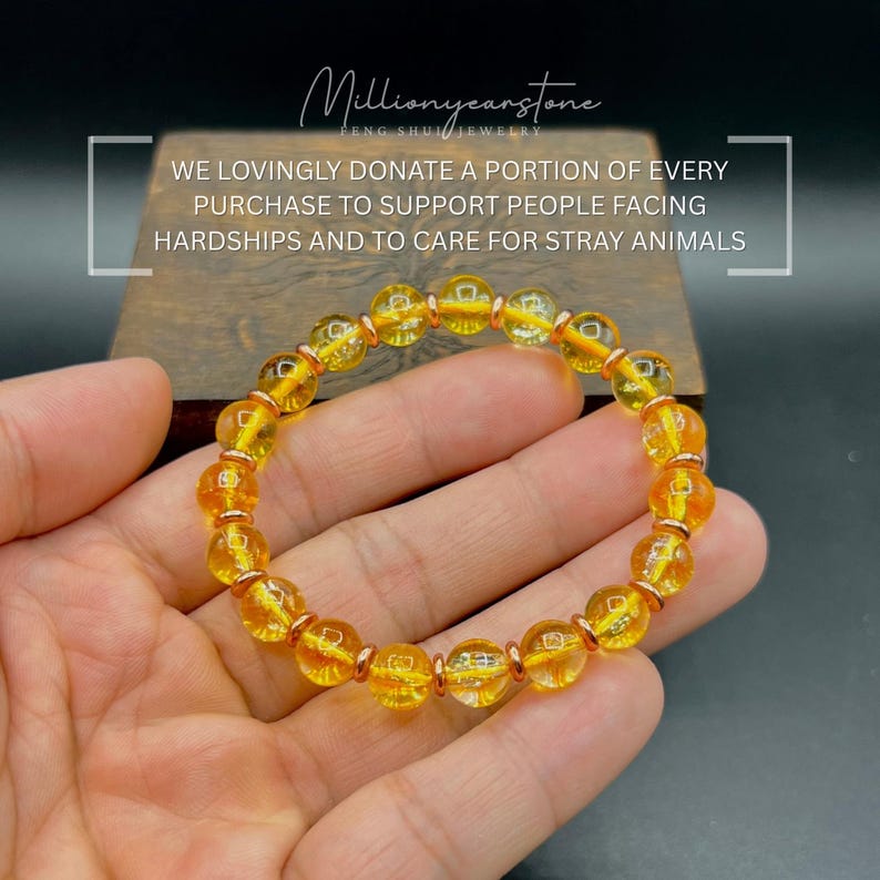 May include: A bracelet of golden yellow beads with copper-coloured spacers. The bracelet is held in a hand, displaying the polished, spherical beads. Text on the image reads: "WE LOVINGLY DONATE A PORTION OF EVERY PURCHASE TO SUPPORT PEOPLE FACING HARDSHIPS AND TO CARE FOR STRAY ANIMALS."