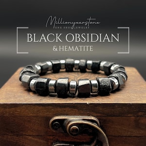 May include: A black obsidian and hematite bracelet. The bracelet features dark, faceted beads and silver-coloured metal spacers. The text "BLACK OBSIDIAN & HEMATITE" is displayed in white, along with the brand name "Millionyearstone". The bracelet rests on a wooden box.