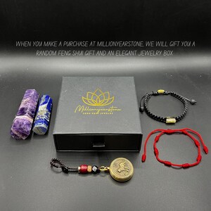Op de afbeelding: Een zwarte doos met een gouden logo met de tekst "Millionyearstone Feng Shui Jewelry". De doos bevat een selectie Feng Shui-sieraden, waaronder een zwarte armband met gouden kralen, een rode armband met gouden kralen, een gouden hanger met een drakenontwerp en twee kristallen staven, een paarse en een blauwe.