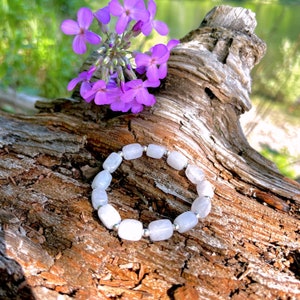 May include: A white gemstone bracelet with silver beads on a wooden background. The bracelet is made of smooth, rounded stones and is a light gray color. The background is a natural wood with a rough texture.