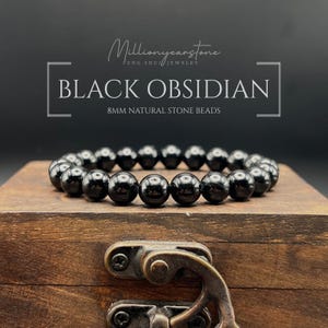May include: A black obsidian bracelet with 8mm natural stone beads. The bracelet is displayed on a wooden box with a metal clasp. The text "BLACK OBSIDIAN" and "8MM NATURAL STONE BEADS" is visible.