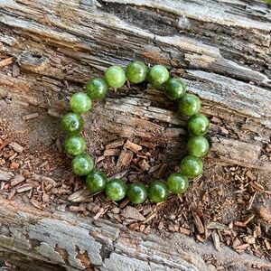 May include: A green beaded bracelet composed of round, polished beads. The bracelet is displayed on a textured, brown wooden surface, creating a natural aesthetic. The beads have a glossy finish.