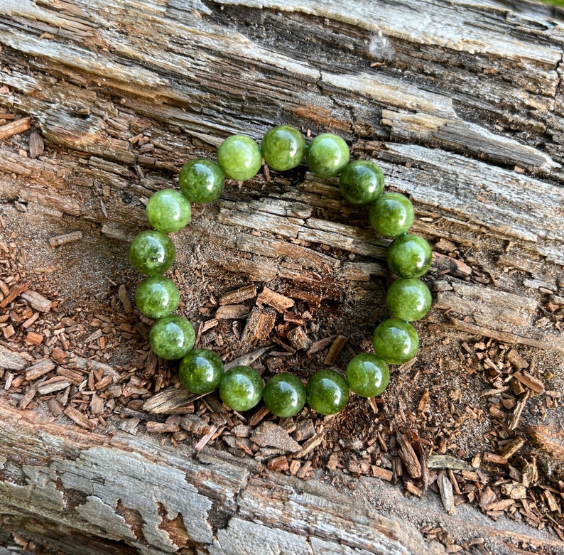 May include: A green beaded bracelet composed of round, polished beads. The bracelet is displayed on a textured, brown wooden surface, creating a natural aesthetic. The beads have a glossy finish.