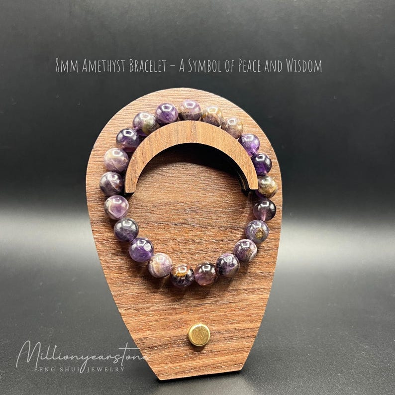 May include: An 8mm amethyst bracelet displayed on a wooden stand. The bracelet is made of purple amethyst beads and is a symbol of peace and wisdom. The text "8MM AMETHYST BRACELET - A SYMBOL OF PEACE AND WISDOM" is printed above the bracelet.