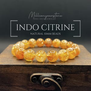 May include: A bracelet of Indo Citrine natural 10mm beads. The beads are a translucent golden-orange color and are strung together. The bracelet is displayed on a wooden box with a metal clasp. The text "INDO CITRINE" and "NATURAL 10MM BEADS" is visible.