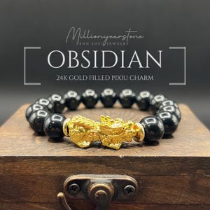 Könnte beinhalten: Obsidian-Armband mit einem 24K goldgefüllten Pixiu-Charm. Das Armband hat schwarze Obsidianperlen und einen goldfarbenen Pixiu-Charm. Der Text "OBSIDIAN" und "24K GOLD FILLED PIXIU CHARM" sind im Bild zu sehen.