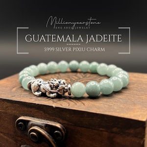 Puede incluir: Un brazalete de jadeíta con cuentas redondas de color verde pálido y un dije Pixiu de plata. El brazalete se exhibe sobre una caja de madera. El texto en la imagen dice "Guatemala Jadeite" y "S999 Silver Pixiu Charm."