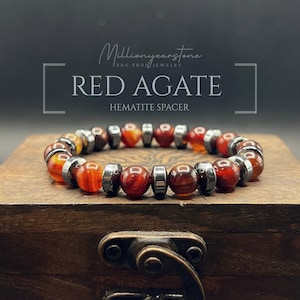 May include: A beaded bracelet featuring red agate and hematite spacers. The bracelet is displayed on a wooden box. The text "Millionyearstone Feng Shui Jewelry" and "RED AGATE HEMATITE SPACER" are visible.