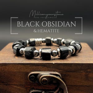 Könnte beinhalten: Armband aus schwarzem Obsidian und Hämatit mit silberfarbenen Akzenten. Das Armband hat facettierte schwarze Perlen und runde silberne Perlen. Der Text "BLACK OBSIDIAN & HEMATITE" ist auf dem Bild zu sehen.