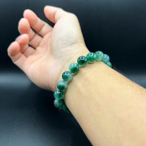 May include: A green jade beaded bracelet with a mottled pattern. The bracelet is worn on a person's wrist.