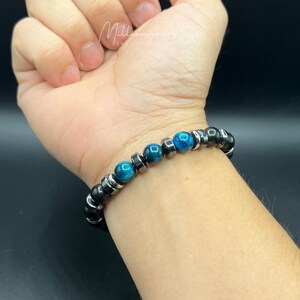 May include: A beaded bracelet featuring black and silver beads, interspersed with vibrant blue tiger eye stones. The bracelet is worn on a wrist, showcasing the contrast of colors and textures. The bracelet is a stylish accessory.