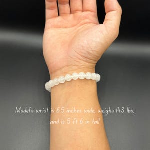 Op de afbeelding: Een witte kralen armband om de pols van een persoon. De armband is gemaakt van kleine, ronde kralen. De pols van de persoon is 6,5 inch breed, weegt 143 pond en is 5 voet 6 inch lang.