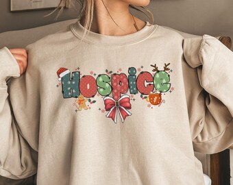 Puede incluir: Una sudadera beige con un diseño navideño que presenta la palabra "Hospice" en letras coloridas con decoraciones festivas como un hombre de jengibre, un lazo y una taza de chocolate caliente.