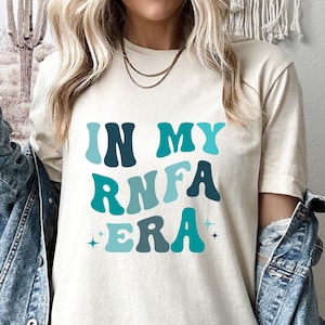 May include: A white t-shirt with the text "IN MY RNA ERA" in blue and teal letters with a retro font.