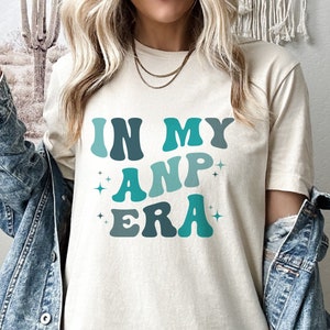May include: A white t-shirt with the text "IN MY AMP ERA" in blue and green letters with a retro font.