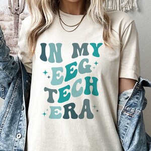Op de afbeelding: Een crèmekleurig t-shirt met de tekst "IN MY EEG TECH ERA" in tinten blauw en turquoise. De tekst is in een retro lettertype met steraccenten. Het shirt is een casual stijl.