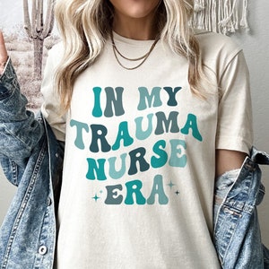 Pu&ograve; includere: Una maglietta bianca con il testo "In My Trauma Nurse Era" in lettere retr&ograve; blu e verdi. Il testo &egrave; circondato da piccole stelle scintillanti.