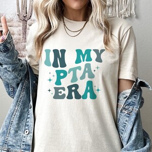 May include: A white t-shirt with the text "In My PTA Era" in blue and green retro-style lettering with sparkling stars.