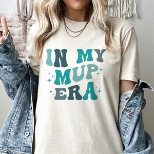以下が含まれることがあります： 白いTシャツに「IN MY MUP ERA」という文字がレトロなフォントで青と緑の文字で印刷されています。文字は白で縁取られており、グラデーション効果があります。テキストの両側に小さな白い星があります。