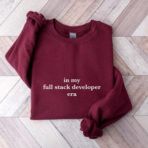 A burgundy sweatshirt with the text "in my full stack developer era" printed on it, laid out on a wooden surface.
