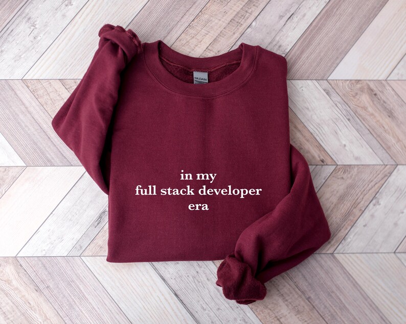 A burgundy sweatshirt with the text "in my full stack developer era" printed on it, laid out on a wooden surface.