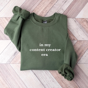 May include: An olive green sweatshirt with the text "in my content creator era" in white. The sweatshirt is made of a soft material and has cuffed sleeves. The garment is displayed on a light wood-grain surface.