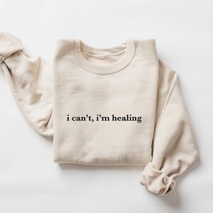 A beige sweater with the text "i can't, I'm healing" printed on it.