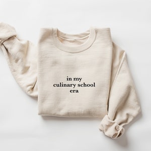 May include: A beige sweatshirt with the text "in my culinary school era" printed on the front.