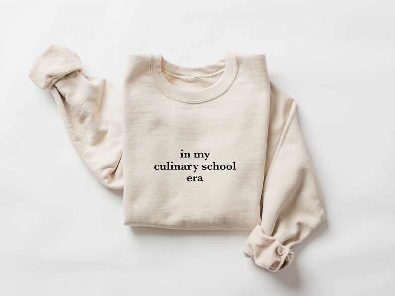 May include: A beige sweatshirt with the text "in my culinary school era" printed on the front.