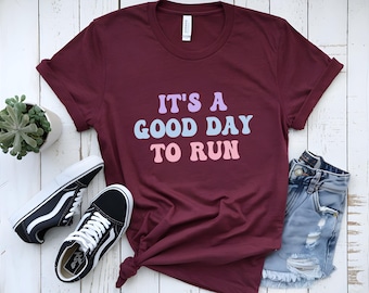 以下が含まれることがあります： パステルカラーの「IT'S A GOOD DAY TO RUN」の文字が入ったバーガンディ色のTシャツ。白と黒のスニーカー、デニムショーツ、小さな鉢植えの多肉植物が白い木製の表面に置かれています。