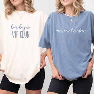 May include: Two oversized t-shirts, one ivory and one blue jean, with short sleeves. The ivory shirt has the text "baby's VIP CLUB" in blue script. The blue jean shirt has the text "mom to be" in white script. Both shirts are paired with black shorts.