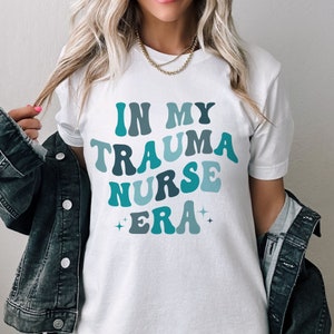 Pu&ograve; includere: Maglietta bianca con il testo "In My Trauma Nurse Era" in lettere blu e turchese con un font retr&ograve;.
