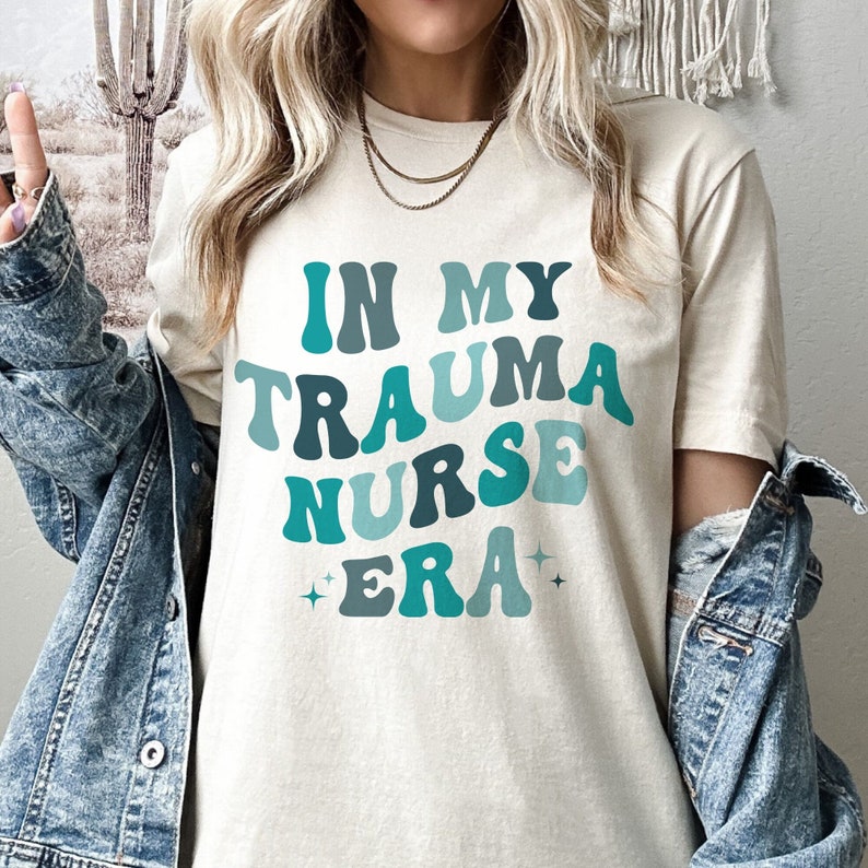 Pu&ograve; includere: Una maglietta bianca con il testo "In My Trauma Nurse Era" in lettere retr&ograve; blu e verdi. Il testo &egrave; circondato da piccole stelle scintillanti.