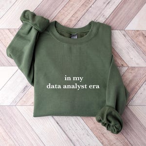 May include: An olive green sweatshirt with the text "in my data analyst era" in white. The sweatshirt is folded neatly on a light brown and white wooden surface. The sleeves are cuffed.