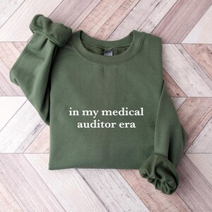 May include: Olive green sweatshirt with the text "in my medical auditor era" in white. The sweatshirt is made of a soft material and has cuffed sleeves. The garment is displayed on a light wood-grain surface.