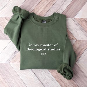 Könnte beinhalten: Ein dunkelgrüner Pullover mit dem Aufdruck "in my master of theological studies era" in weiß auf der Vorderseite.