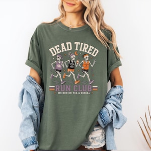 May include: An olive green t-shirt featuring a graphic of three skeletons running. The text reads DEAD TIRED RUN CLUB and WE RUN ON TEA & DENIAL. The skeletons wear athletic gear, headbands, and sneakers while holding cups and a phone.