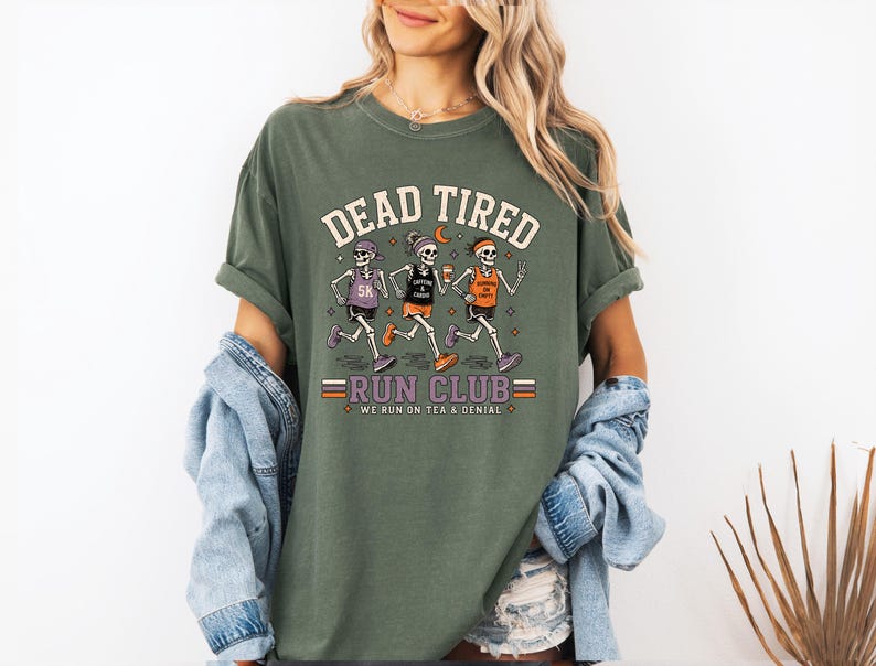 May include: An olive green t-shirt featuring a graphic of three skeletons running. The text reads DEAD TIRED RUN CLUB and WE RUN ON TEA & DENIAL. The skeletons wear athletic gear, headbands, and sneakers while holding cups and a phone.