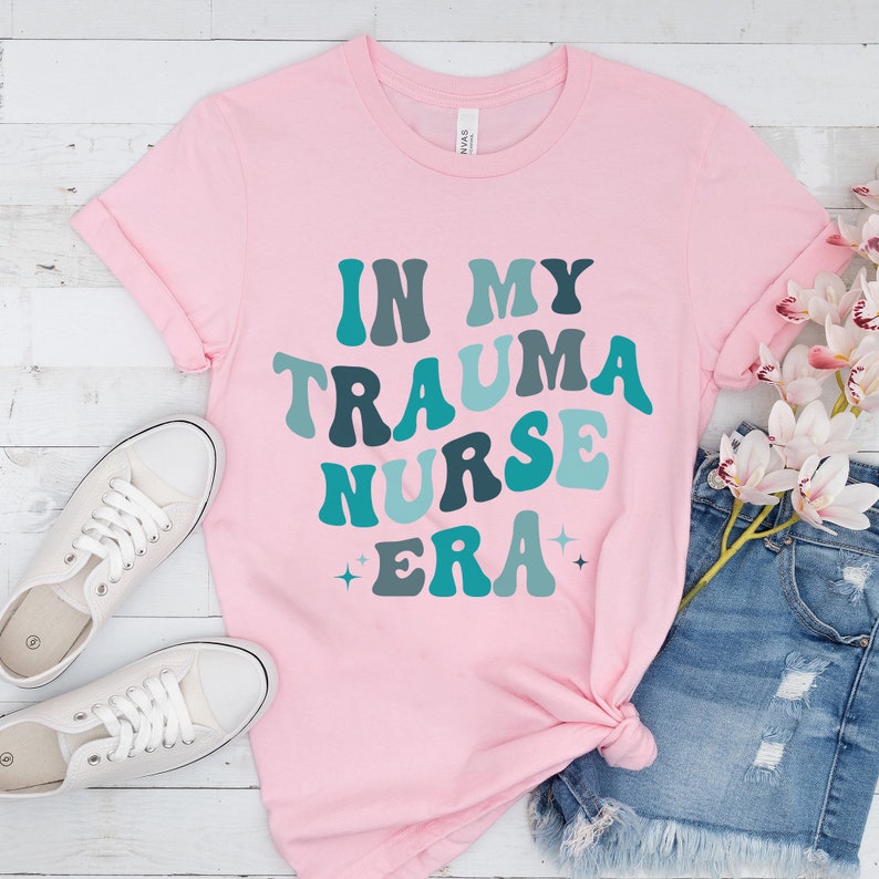 Pu&ograve; includere: Maglietta rosa con il testo "In My Trauma Nurse Era" in un font retr&ograve; con accenti blu e verdi.