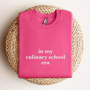 May include: Pink sweatshirt with the text "in my culinary school era" printed in white.