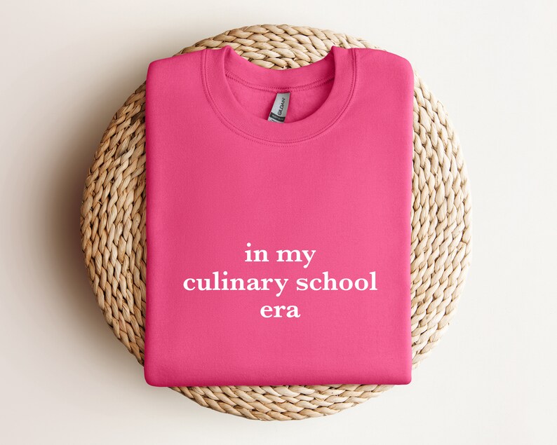 May include: Pink sweatshirt with the text "in my culinary school era" printed in white.