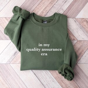 May include: A dark green sweatshirt with the text "in my quality assurance era" printed in white on the front.
