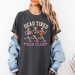 May include: A charcoal gray t-shirt featuring a graphic of three skeletons running. The text reads DEAD TIRED RUN CLUB. The skeletons wear athletic gear with text including 13.1, Last Mile, and Rest in Pace. The design uses orange, purple, and white.