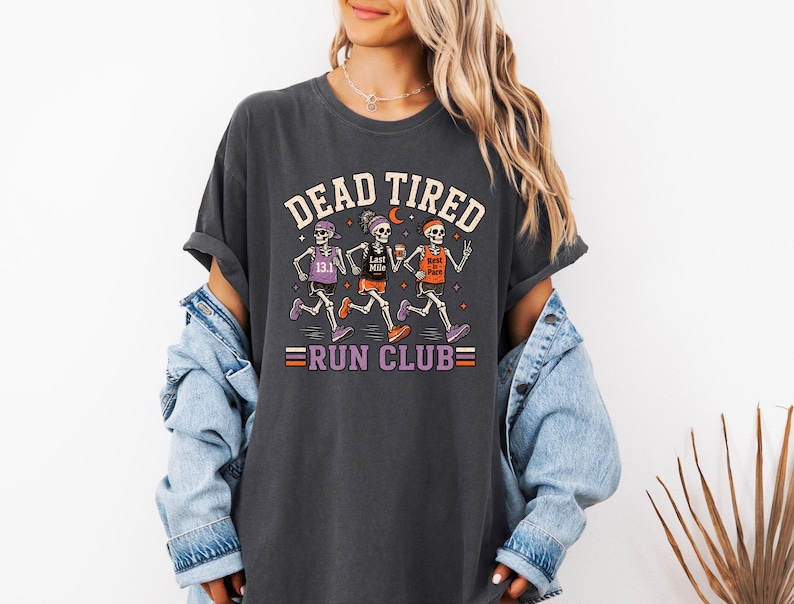May include: A charcoal gray t-shirt featuring a graphic of three skeletons running. The text reads DEAD TIRED RUN CLUB. The skeletons wear athletic gear with text including 13.1, Last Mile, and Rest in Pace. The design uses orange, purple, and white.