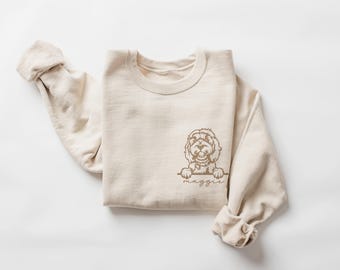 May include: A cream-colored sweatshirt with a crew neck and long sleeves. A brown illustration of a dog peeking over a line is on the front. The dog's name, "Maggie", is written below the line in a script font.