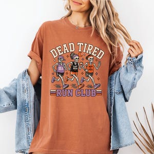 May include: A rust-colored short-sleeve t-shirt featuring a graphic of three skeletons running. The text reads DEAD TIRED RUN CLUB. The skeletons wear athletic gear with text including 13.1, Last Mile, and Best in Pace. The design uses orange, purple, and white.